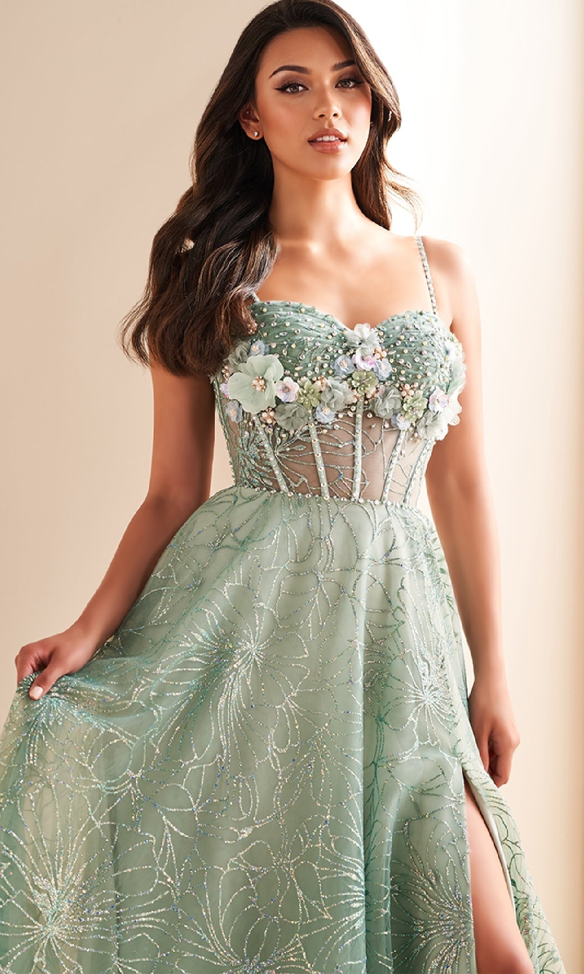 A woman stands indoors against a neutral background wearing the Ellie Wilde EW36218 long prom dress—a light green A-line gown with 3D flowers, sheer corset bodice, thin straps, and a skirt featuring a slit.